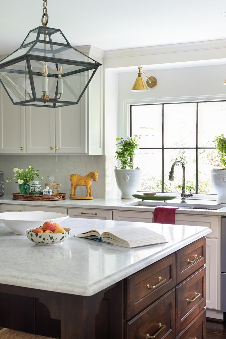 Kitchen island with marble top and walnut cabinetry standing before a deep kitchen sink workstation backed by a large window.