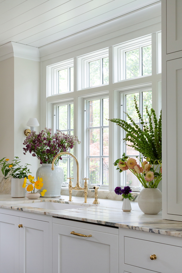 A natural stone countertop fitted with undermount sink and polished brass fixtures sits below a v-groove paneled ceiling. A large paned window is set into the wall behind the sink, flanked by two sconces. 