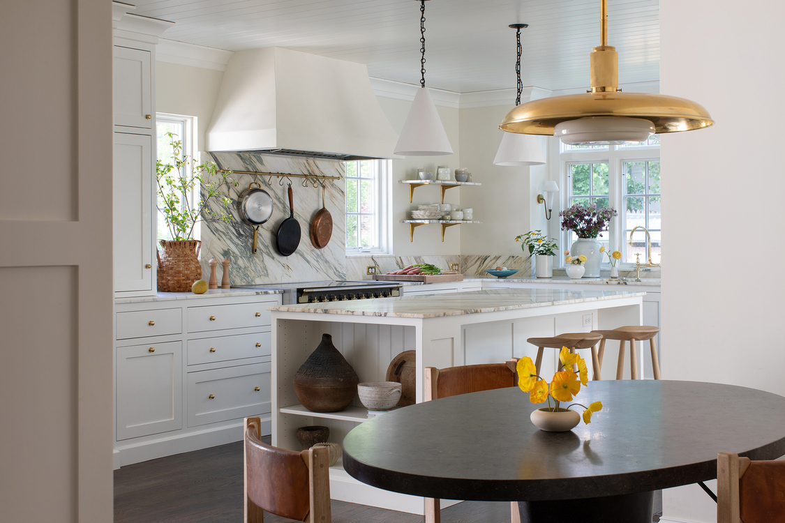 Combination kitchen and dining area with stone slab counters that continue up the wall to the range hood. A custom island with v-groove paneling and inset shelving sits in the center, flanked by the range and sink. Two styles of architectural pendant lighting bring both balance and interest to the space. 