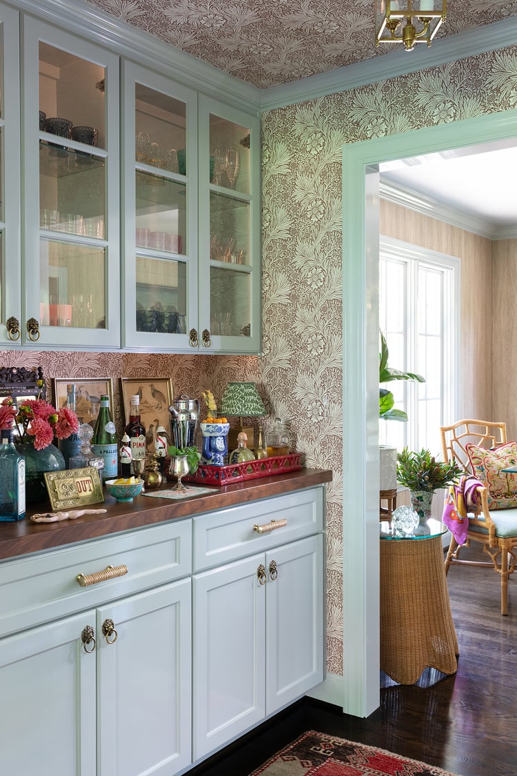 Eclectically decorated kitchen bar with walnut bartop, pale blue cabinetry, Morris & Co wallpaper, lion door-knocker cabinet pulls and woven rattan drawer pulls.