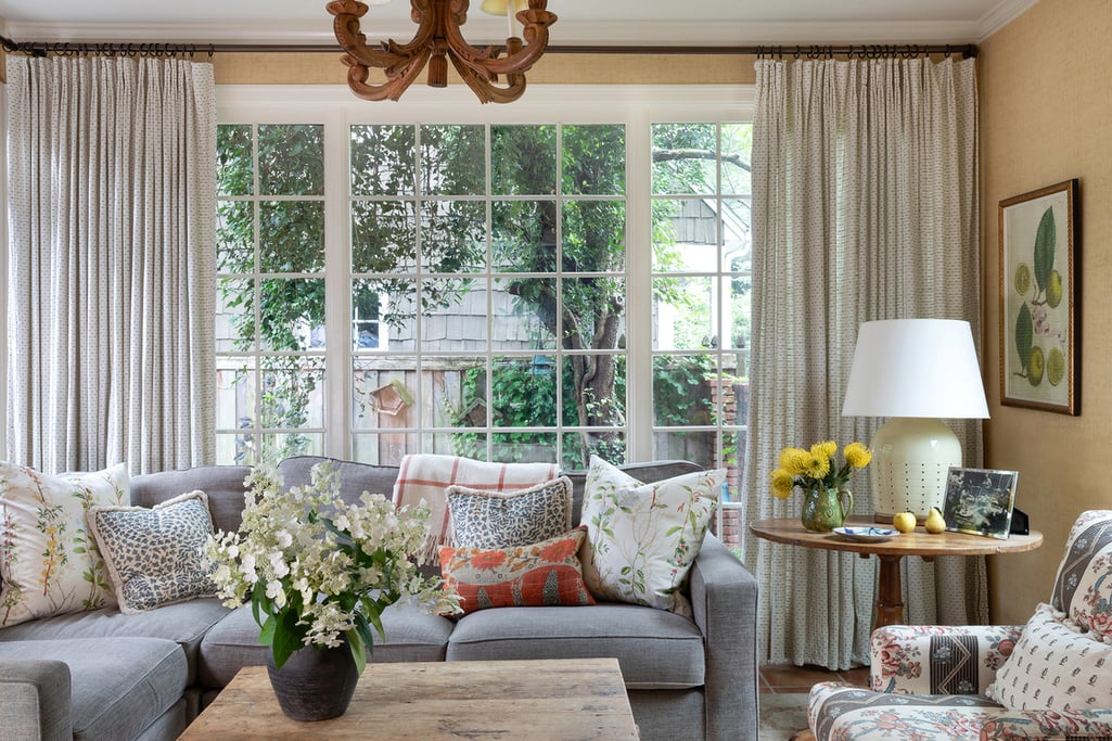 Cozy living space with large window flanked by floor-to-ceiling curtains. Warm-toned grasscloth wallpaper and a carved wood chandelier bring out the red and yellow tones amidst the cool-toned decor..